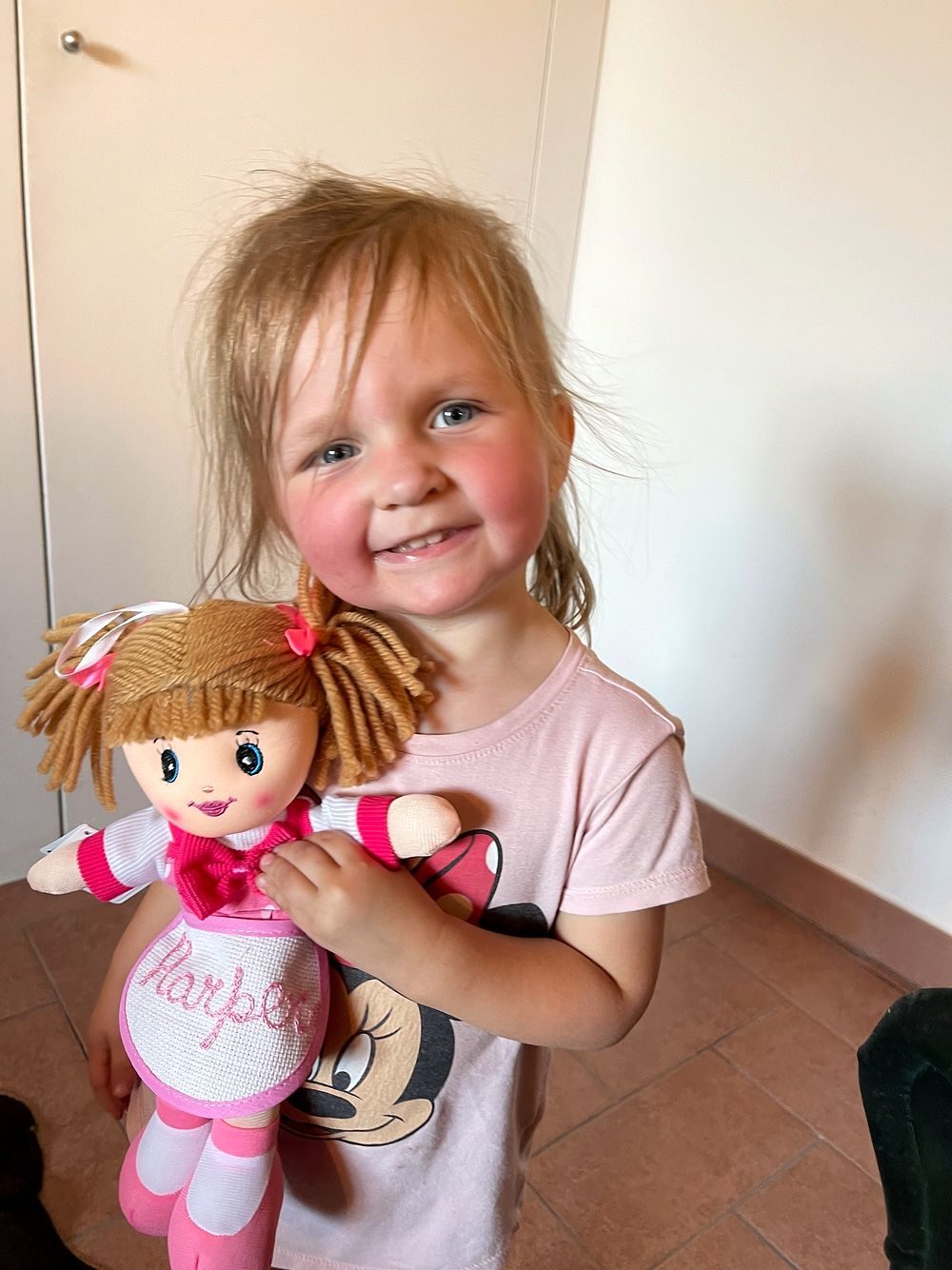 A young blonde girl grins broadly while holding a rag doll with 'Harper' embroidered on its dress, standing on terracotta tiles in a Tuscan apartment