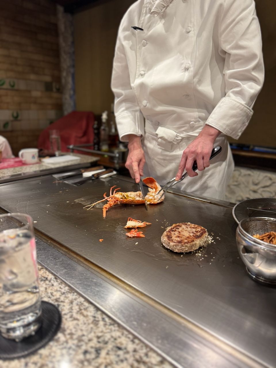 A chef in a white coat works a teppanyaki grill, cutting a whole lobster beside a seared wagyu beef patty, with a mesh strainer and water glass visible on the counter.
