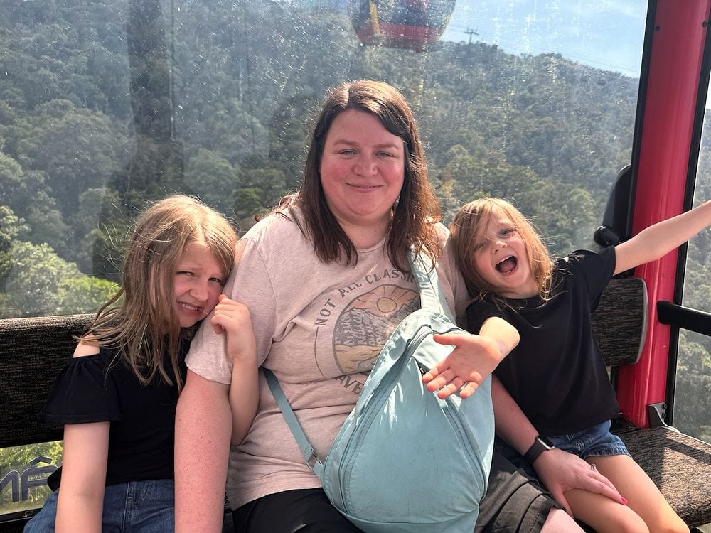 Lindsay seated in a Ba Na Hills cable car gondola with two of her daughters, forested mountains and a second gondola visible through the glass