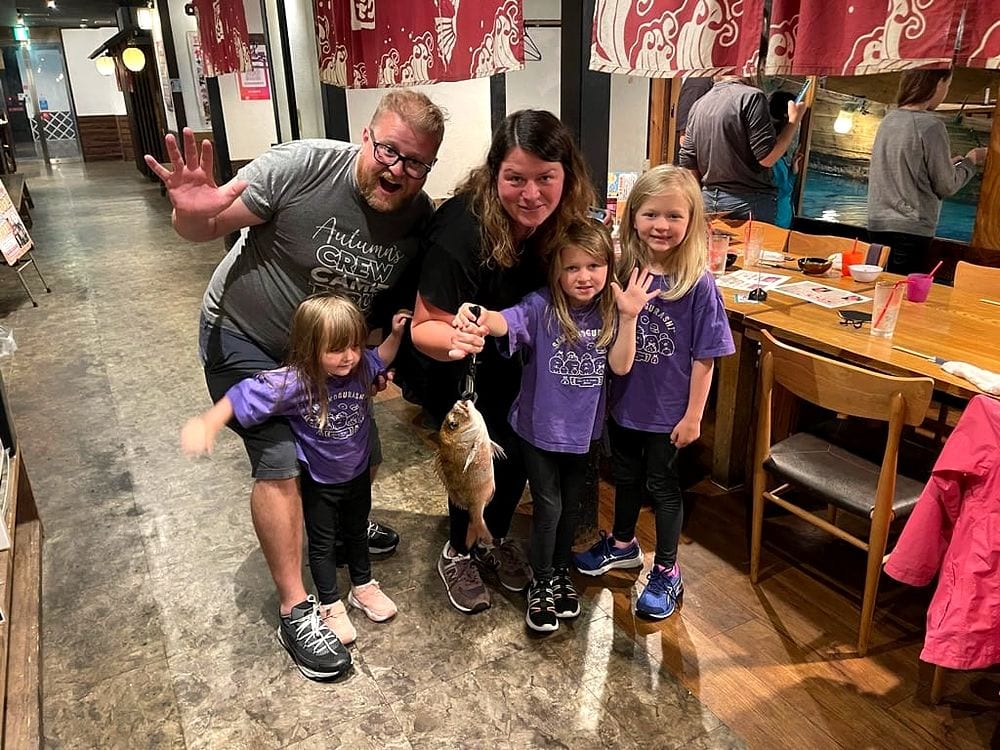 A family of five poses inside Zauo Fishing Restaurant in Namba, Osaka, with one of the girls holding up a whole raw fish on a hook, the group in matching purple shirts.