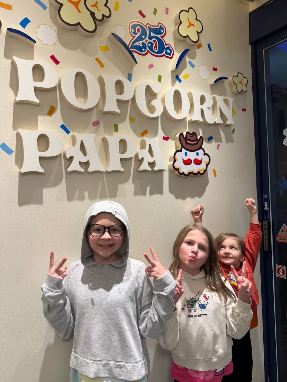 Three girls pose with peace signs and raised fingers in front of a large 3D Popcorn Papa sign with confetti decorations marking the shop's 25th anniversary, the middle girl wearing a Hello Kitty sweatshirt.