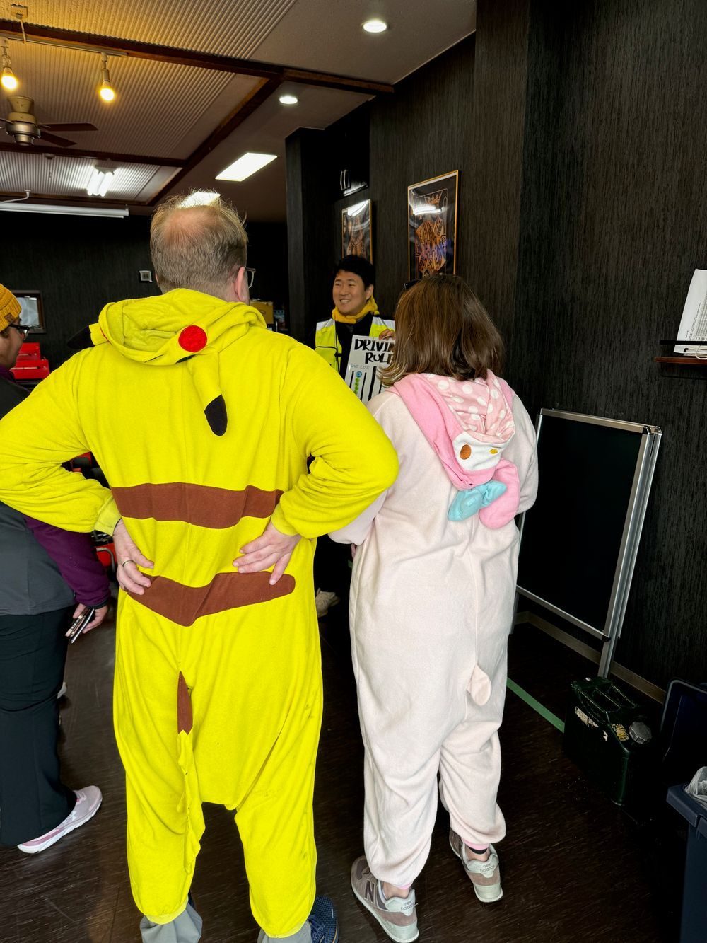 Adam in a Pikachu onesie and Lindsay in a My Melody onesie stand with their backs to the camera, listening to a staff member give a driving rules briefing inside the Street Kart facility in Asakusa.