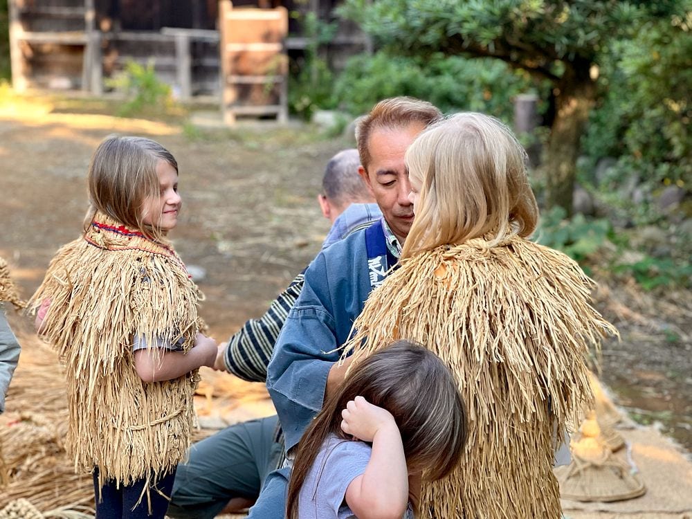 A family in straw hats and capes gathers together smiling outside a historic thatched-roof building at an open-air folk museum.