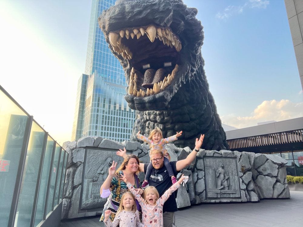 A family of five poses in front of the large Godzilla statue at Kabukicho Tower in Shinjuku, with the parents and three young girls throwing their arms up and making excited faces, the youngest child perched on the dad's shoulders.