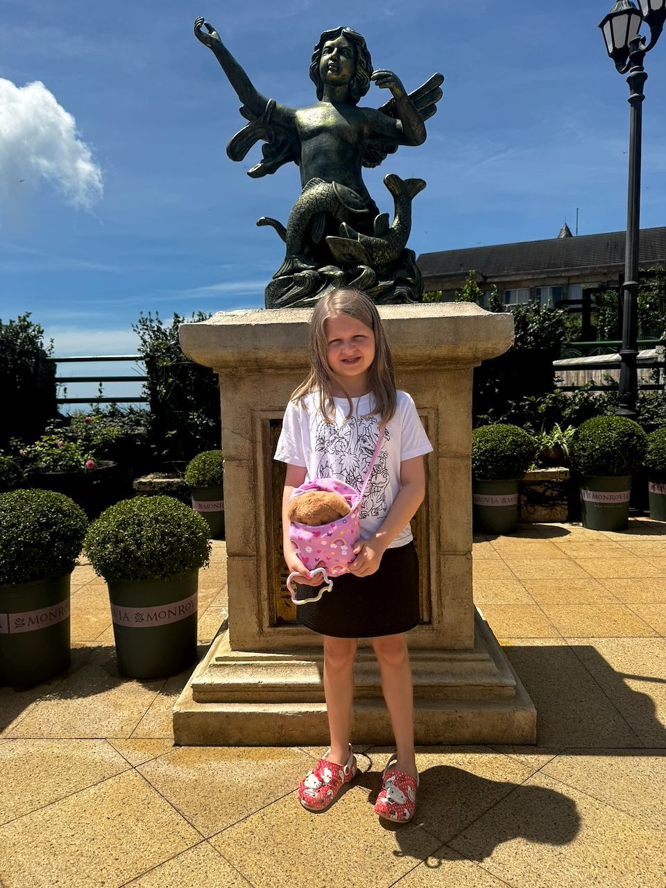 A young girl holding a stuffed animal stands in front of a bronze mermaid statue on a sunlit hotel terrace at Ba Na Hills