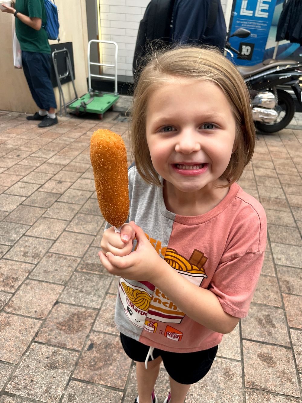 A young girl on a Seoul sidewalk holding up a golden fried Korean corn dog with both hands and a huge smile