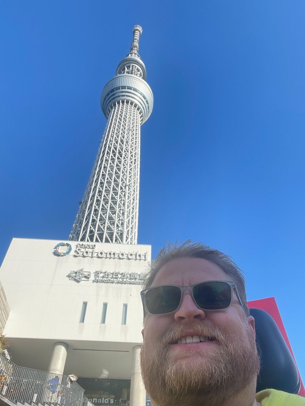 A man in sunglasses takes a selfie from a low angle in front of the Tokyo Skytree and the Solamachi shopping complex on a clear blue-sky day.