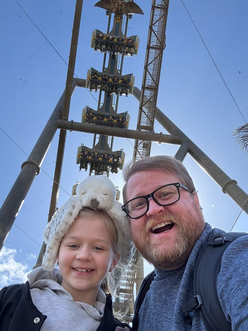 Adam and Lily take a selfie in front of a tall hanging roller coaster at Universal Studios Japan, with Lily wearing a fluffy owl hat and riders visible in the suspended coaster cars high above them.