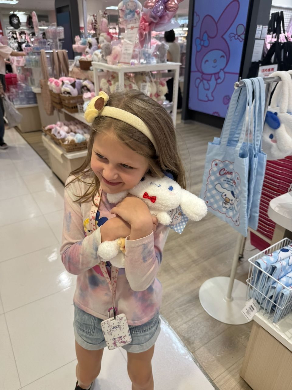 Cora stands in a Sanrio store in Ginza hugging a Cinnamoroll plush toy, wearing a Sanrio headband and a floral lanyard, with shelves of Sanrio merchandise visible behind her.