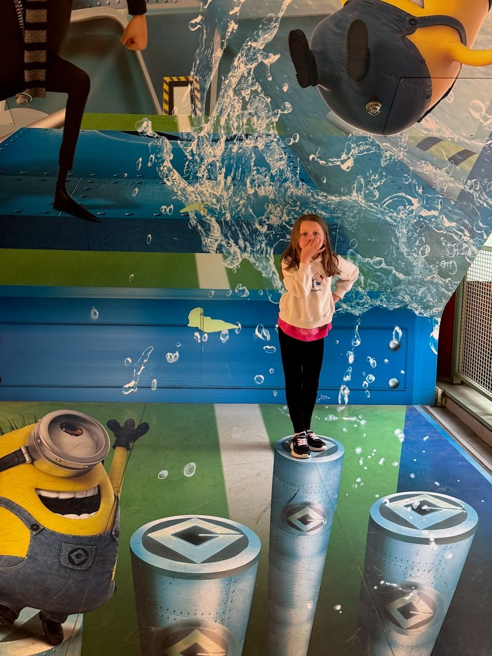 A young girl stands on a painted 3D cylindrical pillar covering her mouth in mock surprise, surrounded by a trompe-l'oeil mural of splashing water, a laughing Minion, and Gru at Universal Studios Japan.