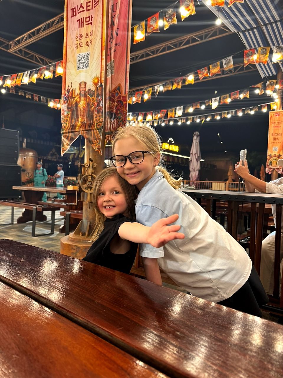 Two young girls sit together at a long wooden table inside a festively lit outdoor venue with Korean-language banners and string lights overhead, the older one leaning over to hug the younger one who grins at the camera.