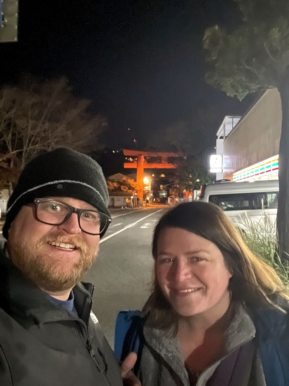 A man and a woman take a nighttime selfie on a street, with a large illuminated red torii gate visible in the background down the road.