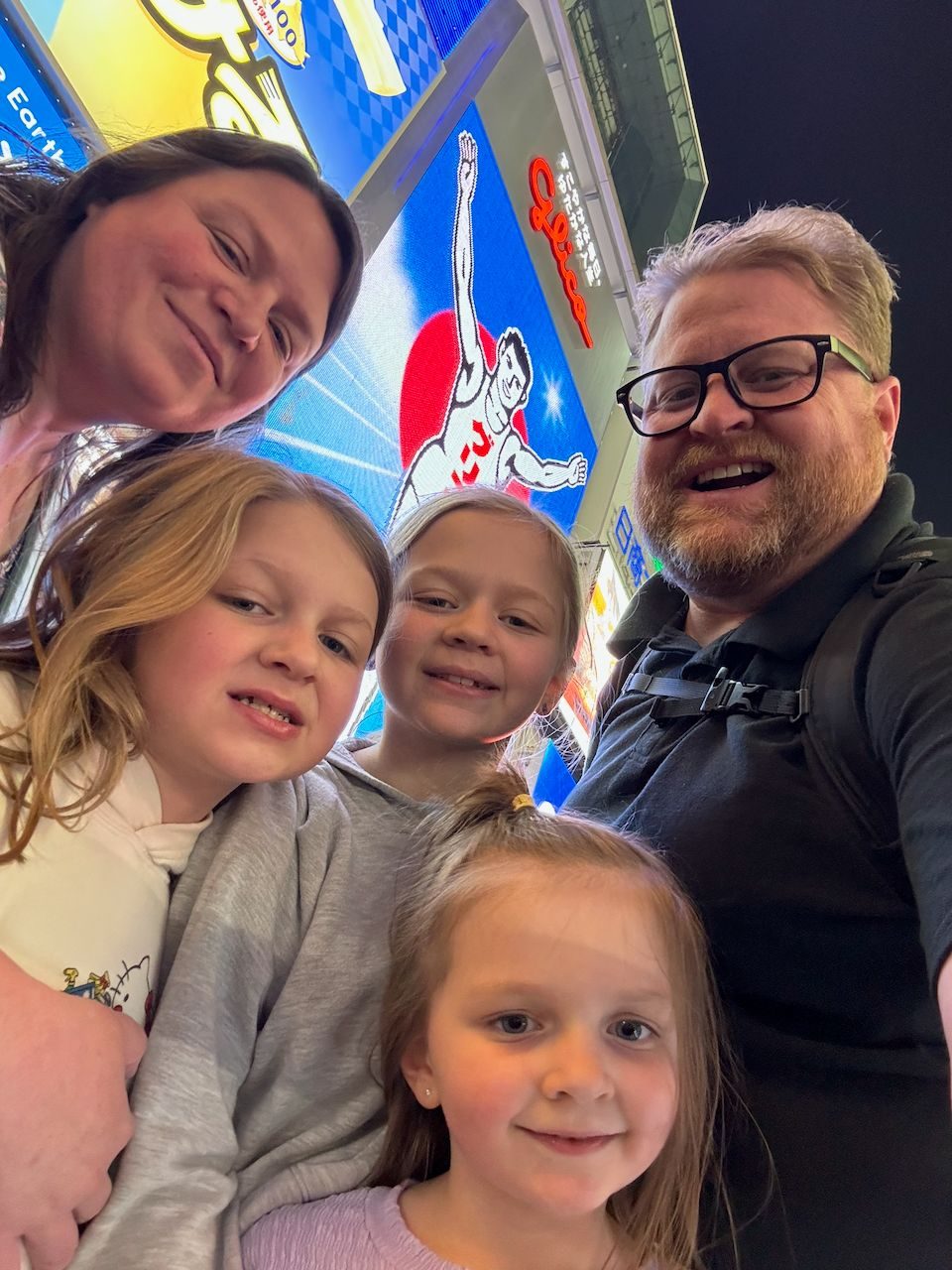 Family selfie of Adam, Lindsay, and their three daughters at night on Dotonbori with the Glico Man sign lit up behind them