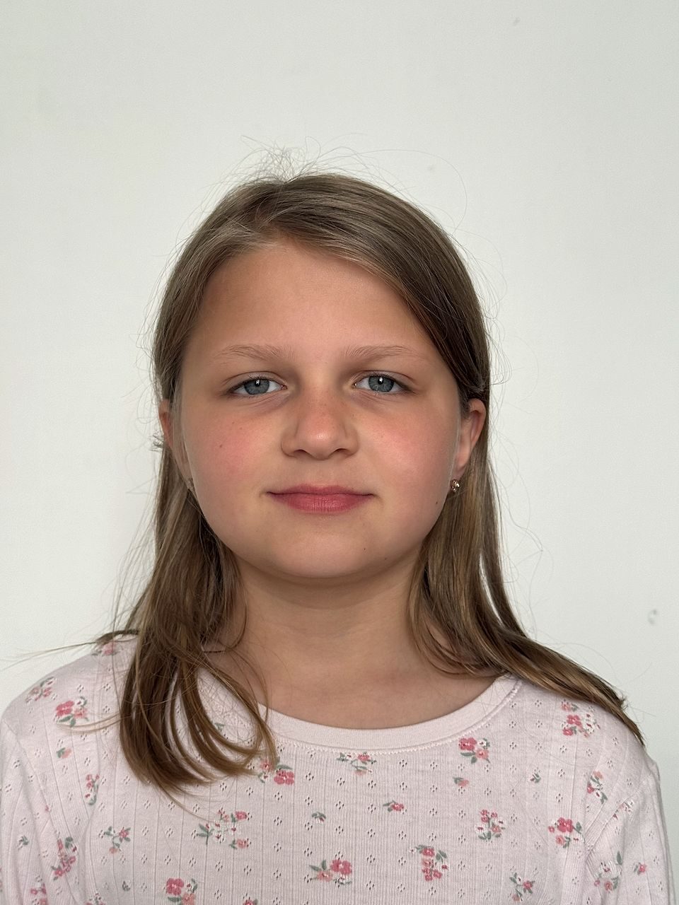 A young girl with blue eyes and long brown hair wearing a light pink floral top looks directly at the camera against a plain white background in a passport-style photo