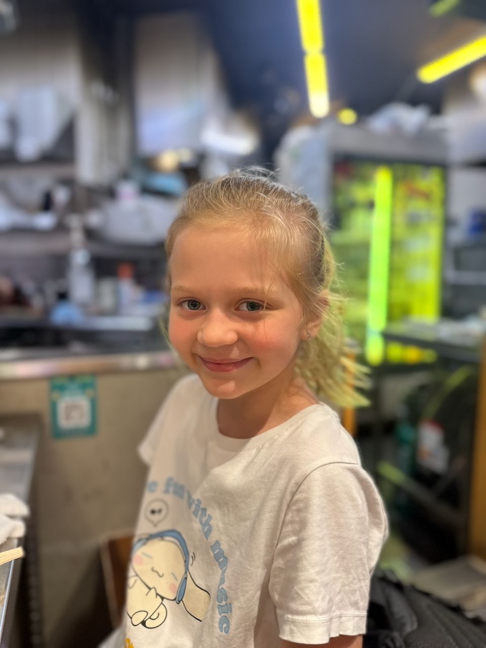 A young blonde girl smiles over her shoulder at the camera while seated at a restaurant counter, with a busy open kitchen visible in the background.