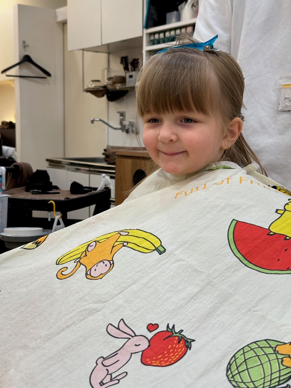 A young girl smiles while wearing a colorful cutting cape printed with fruit patterns including bananas, watermelon, and strawberries.