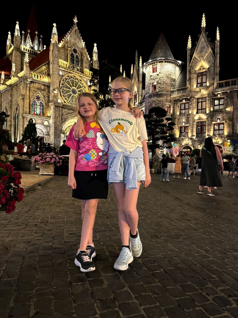 Two girls stand arm in arm on a cobblestone plaza at night in front of illuminated Gothic-style castle and cathedral buildings at Ba Na Hills, one wearing a pink Hello Kitty shirt and the other a cream Cinnamoroll tee.