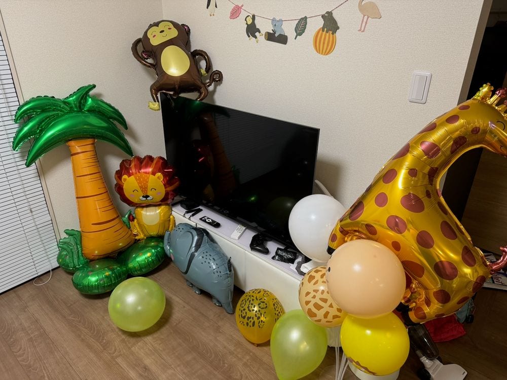 A corner of a Japanese apartment living room decorated with safari-themed foil balloons including a giraffe, lion, elephant, monkey, and palm tree, surrounded by latex balloons in green, yellow, and white, with an animal garland strung on the wall above a flat-screen TV.