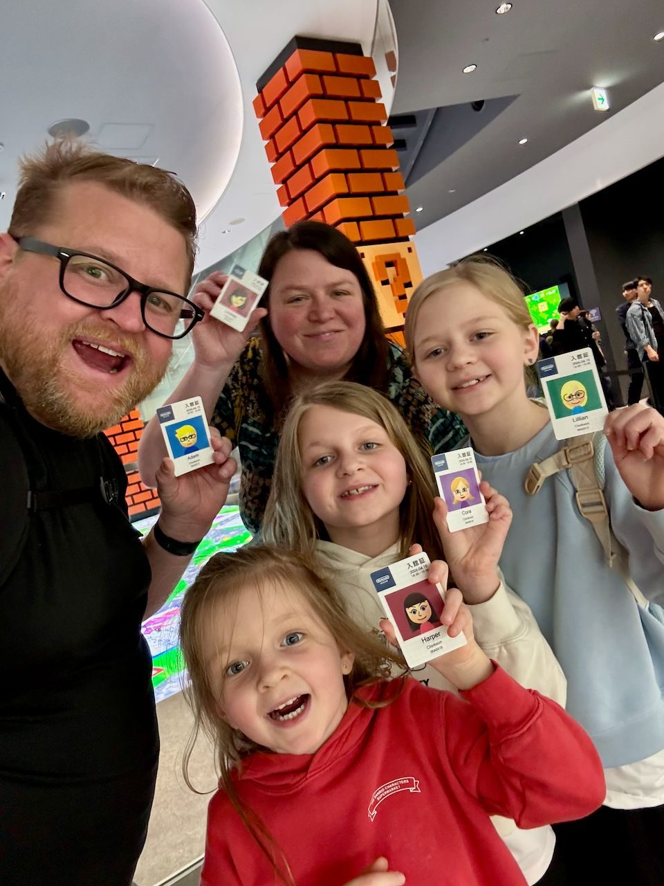 A family of five takes a selfie inside Super Nintendo World, each holding up their personalized Mii entry cards, with a giant Mario brick column visible in the background.
