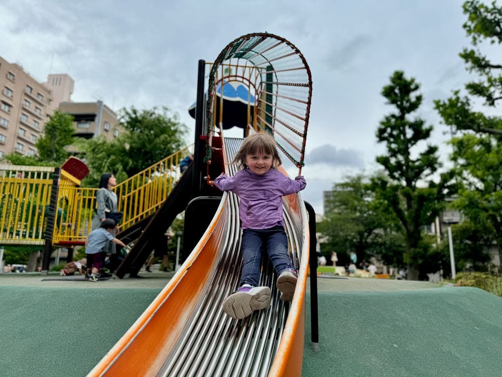 A young girl slides down an orange slide at Iriya Minami Park, emerging from a caged climbing tunnel structure at the top with yellow playground equipment and city buildings visible behind her.