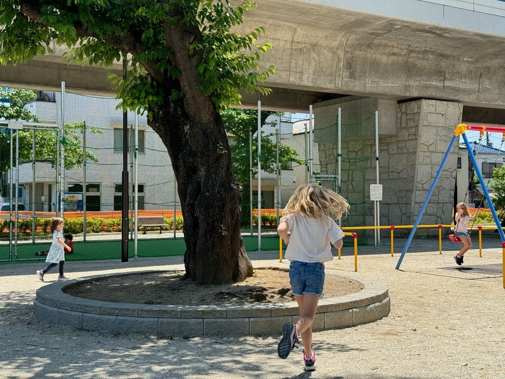 A girl runs past a large tree toward a swing set built into the space underneath an elevated train line in a Tokyo neighborhood.