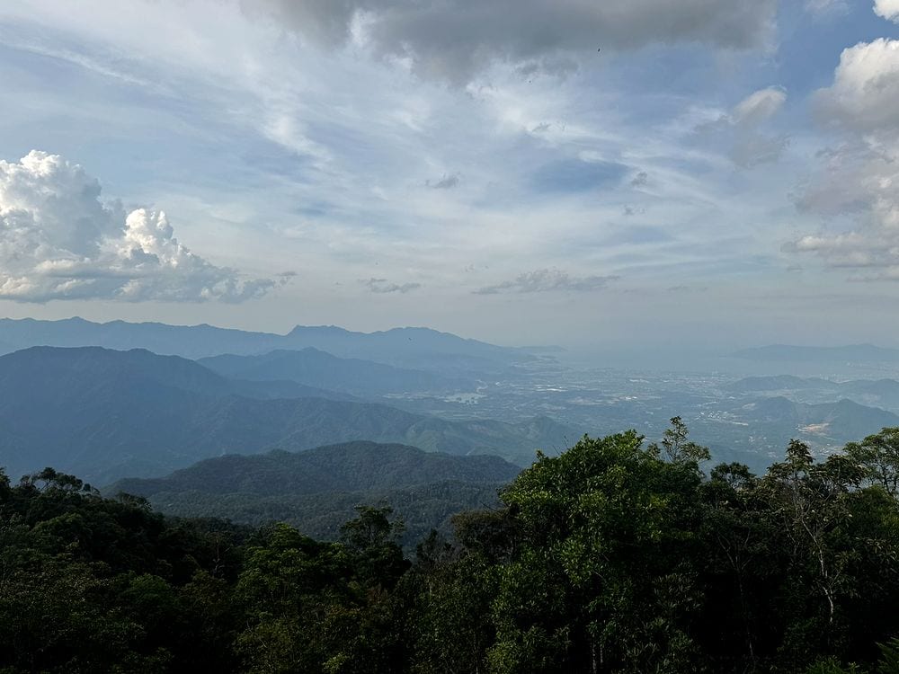 A wide view from Ba Na Hills looking out over layered forested mountain ridges fading into haze, with the Da Nang valley visible in the far distance under a partly cloudy sky.