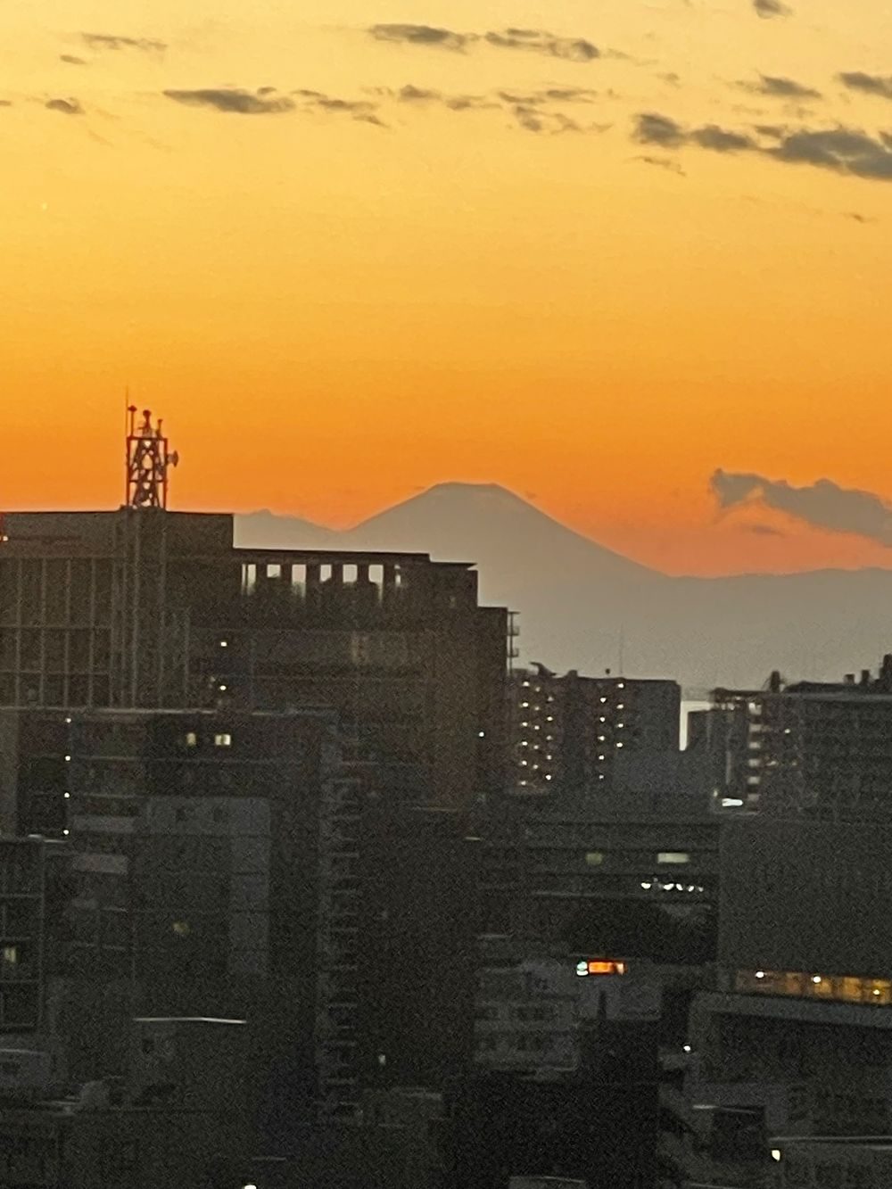 Mount Fuji's silhouette rises above a Tokyo-area skyline at sunset, the sky glowing deep orange behind city buildings.