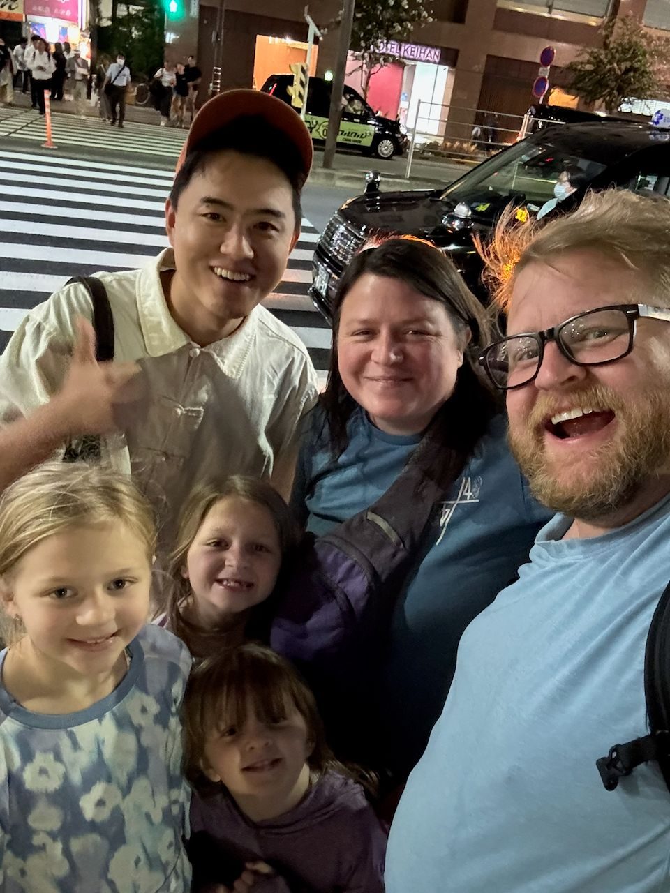 A nighttime selfie on a busy Asakusa street corner showing a family of five with a young male subscriber who is waving at the camera, with a lit crosswalk and taxi visible behind them.