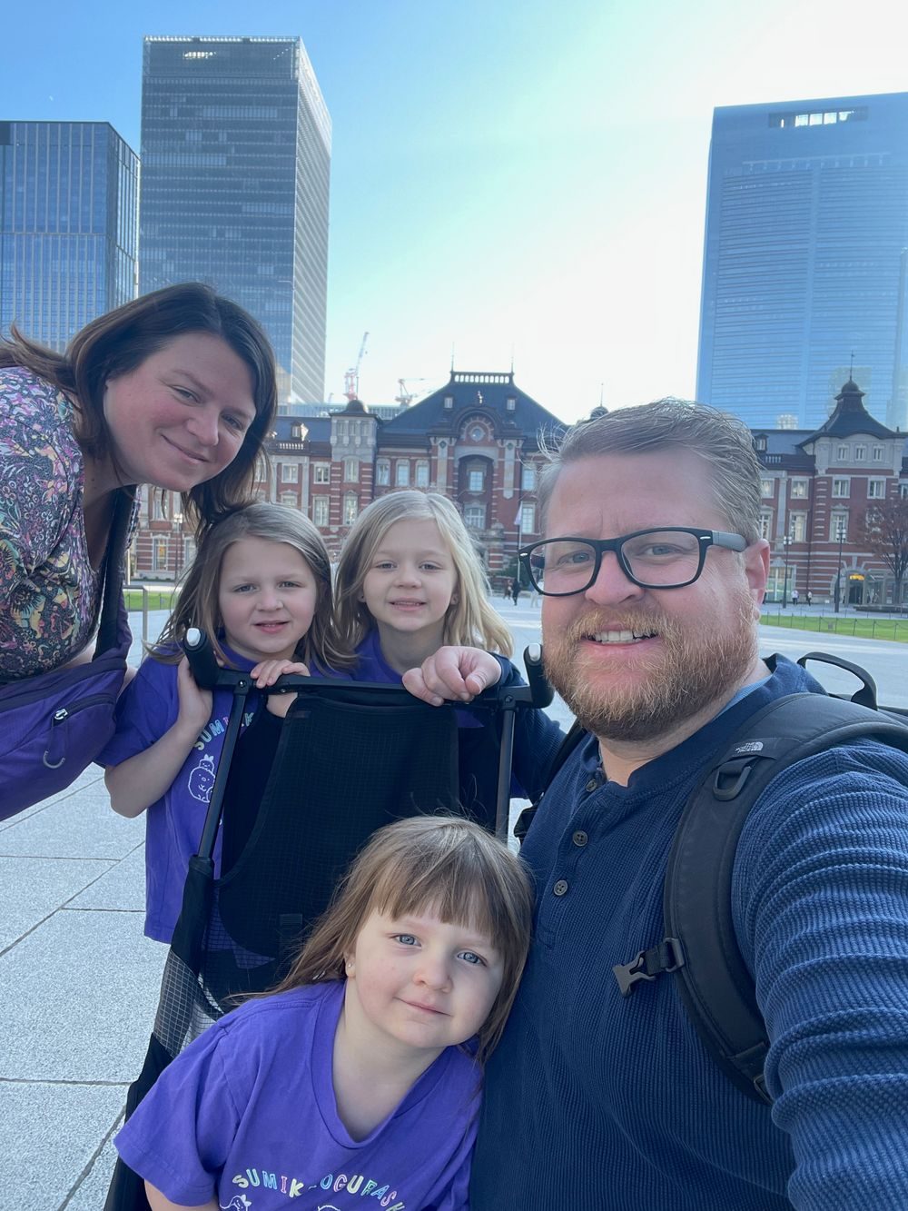 A family of five poses in front of the red brick Marunouchi facade of Tokyo Station on a sunny day, with modern office towers visible behind them.