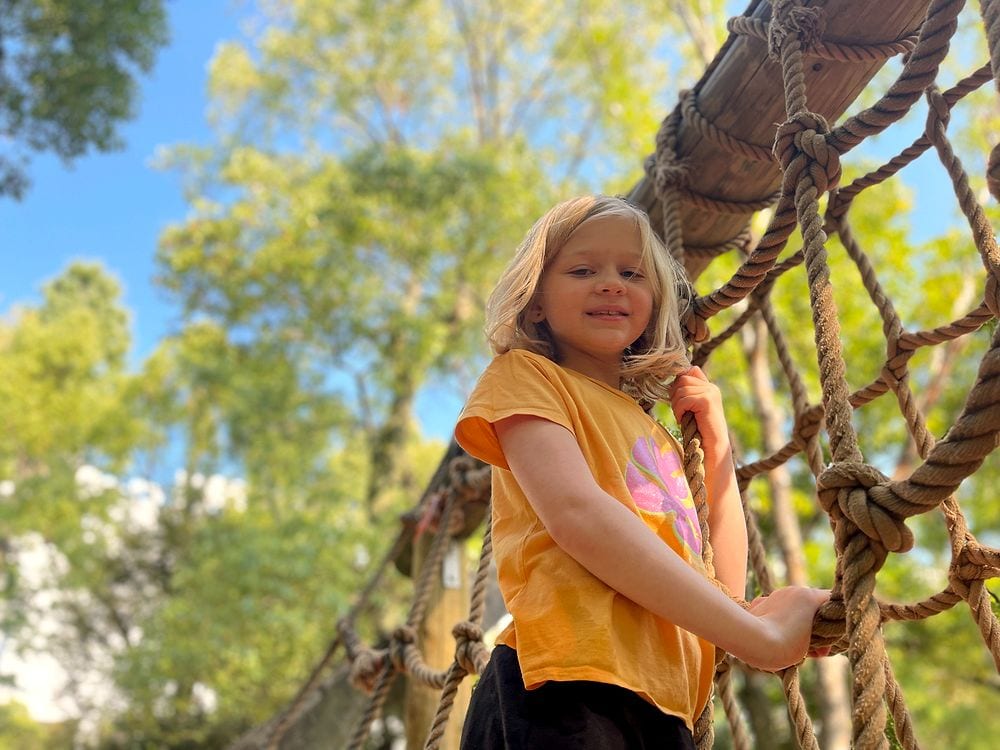 A young girl stands on a rope climbing structure at Heiwa no Mori Park in Ota, Tokyo, smiling at the camera with trees in the background.
