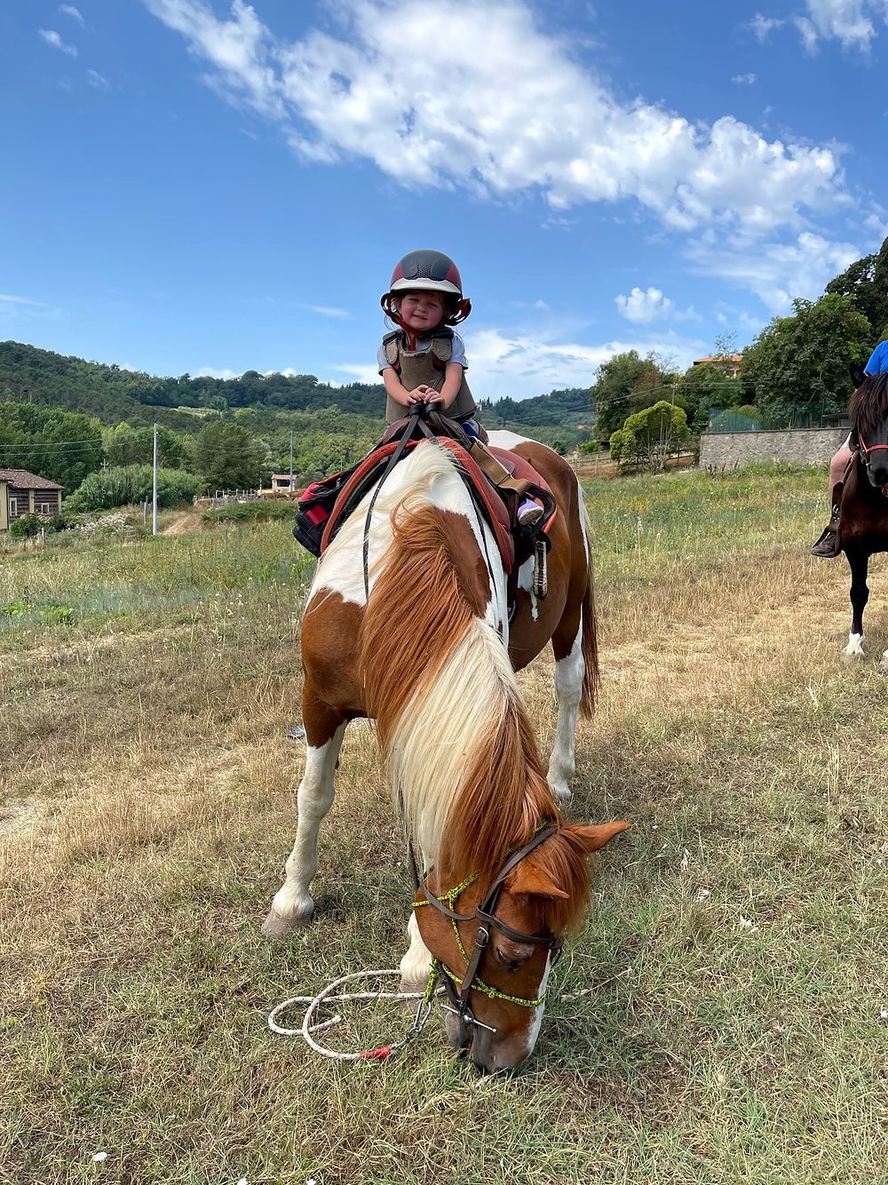 A young child in a riding helmet and protective vest sits on a brown and white pinto horse that is grazing in a dry Tuscan field, with green hills and blue sky behind them