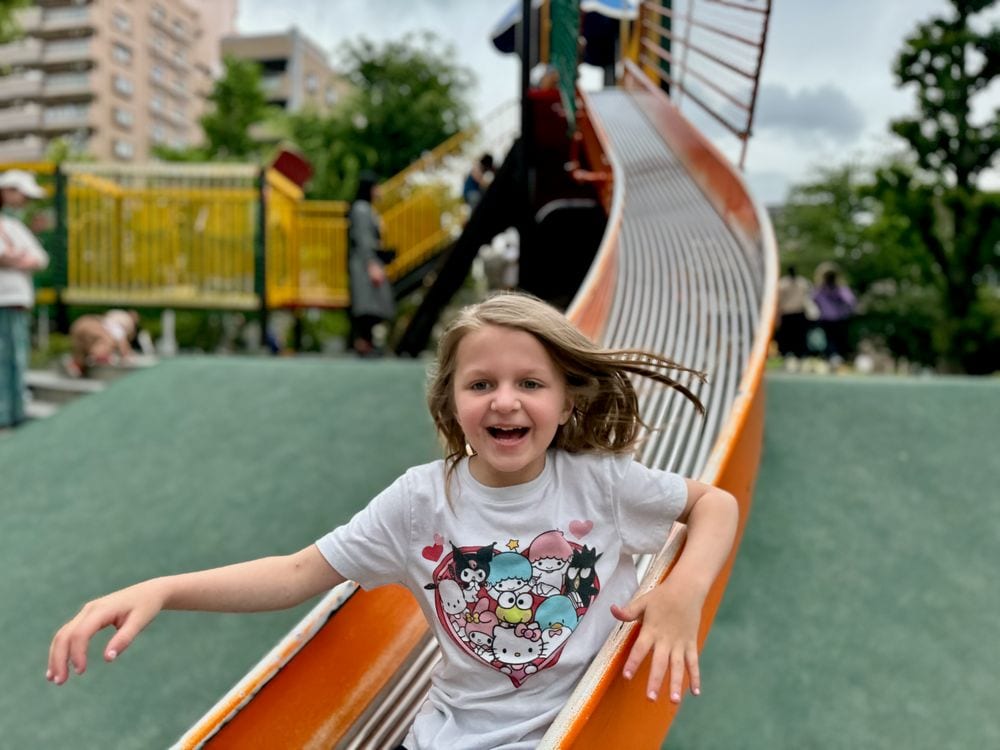 A young girl wearing a Sanrio characters t-shirt slides down a long orange playground slide with her mouth open in delight, hair flying, at an outdoor park in Tokyo.